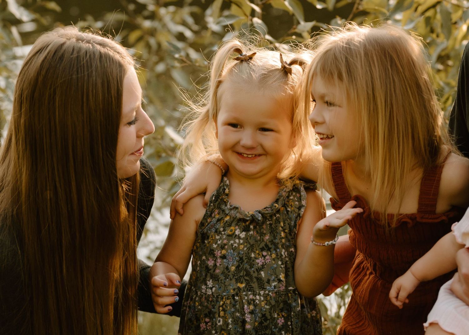 A mother and two young daughters share a joyful moment outdoors in warm golden light.