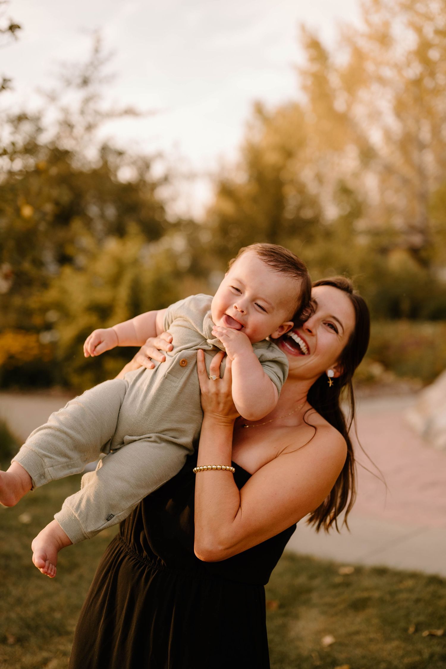Laughing mother lifting giggling baby boy outdoors in warm golden hour autumn light.