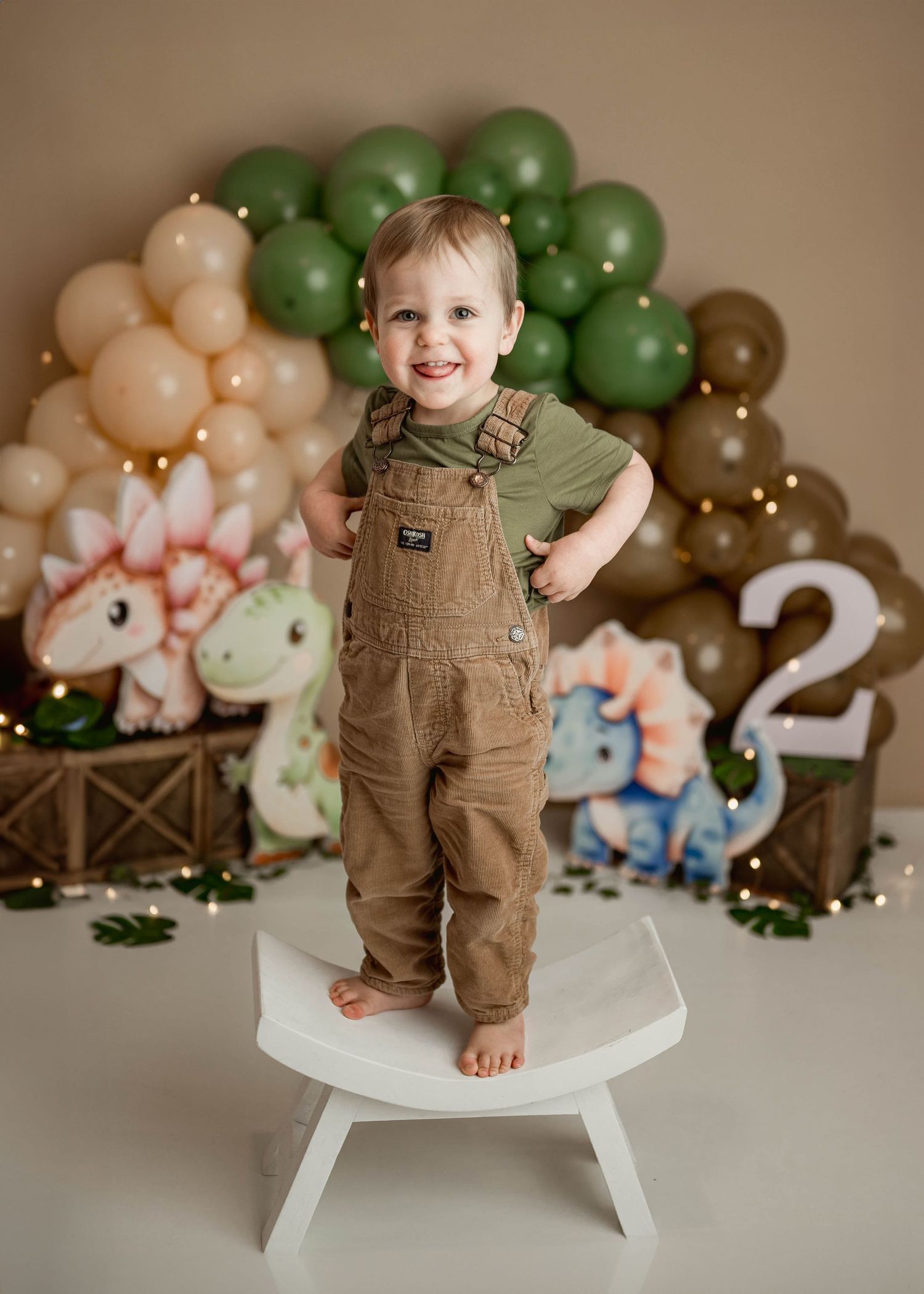 Smiling toddler boy in overalls standing on white stool with dinosaur themed green balloon arch backdrop.