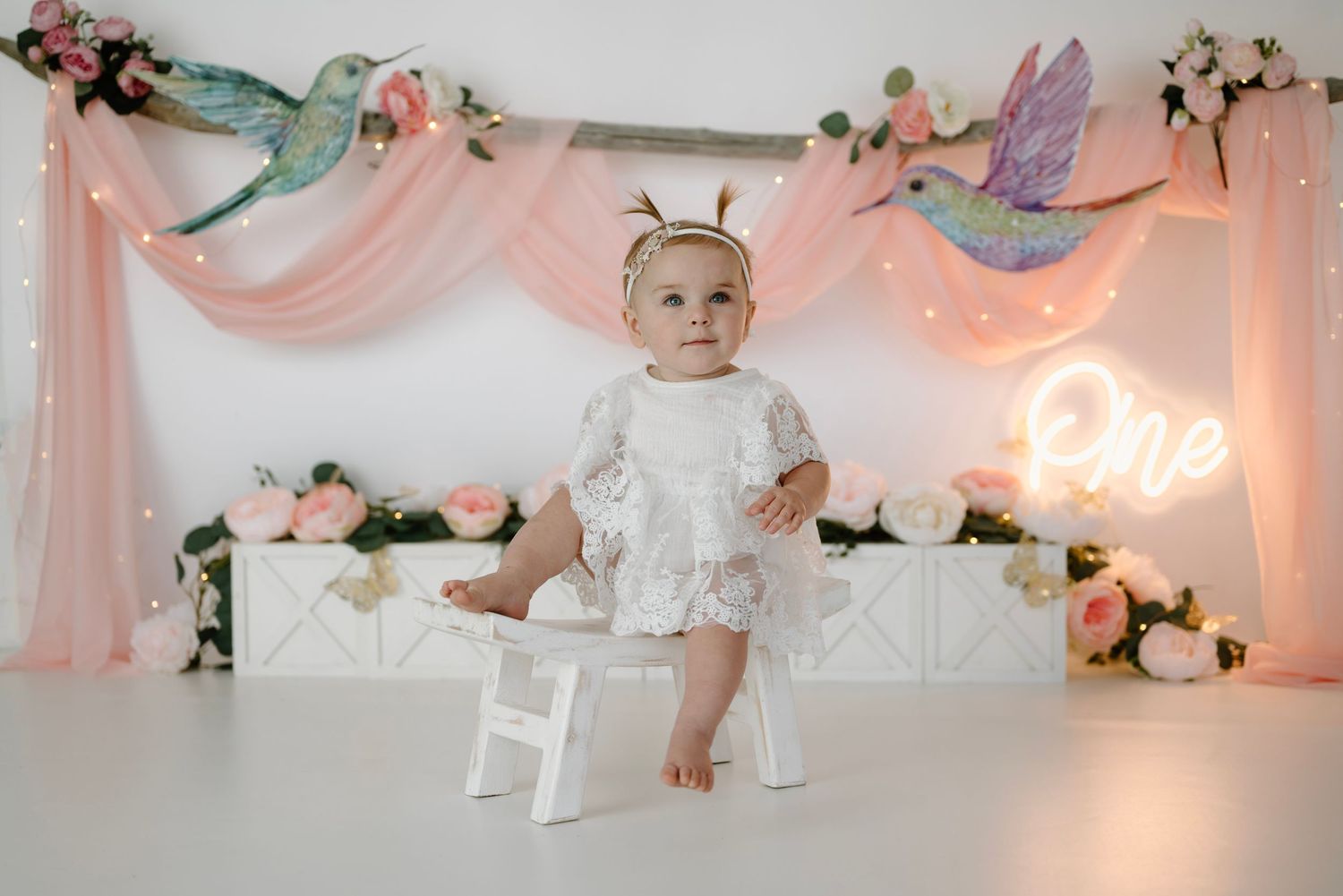 Baby girl in white dress stands on white stool at a pink floral hummingbird-themed first birthday setup.
