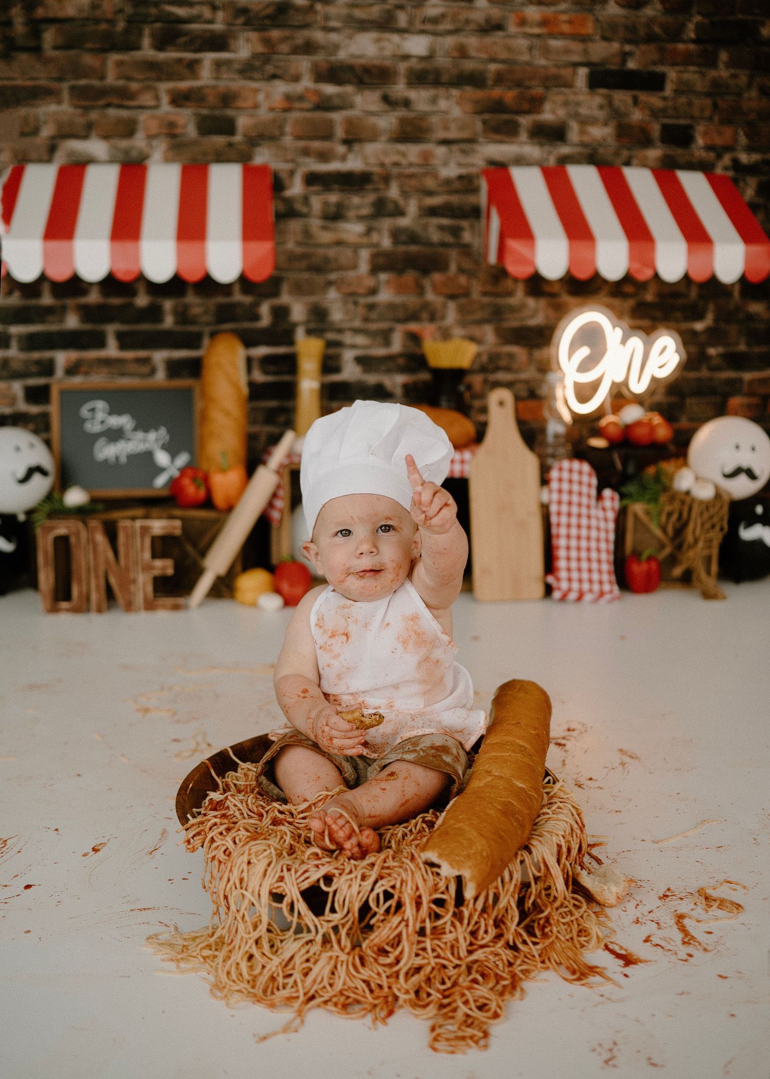 Baby in chef hat waves while sitting in spaghetti nest with baguette at Italian first birthday shoot.