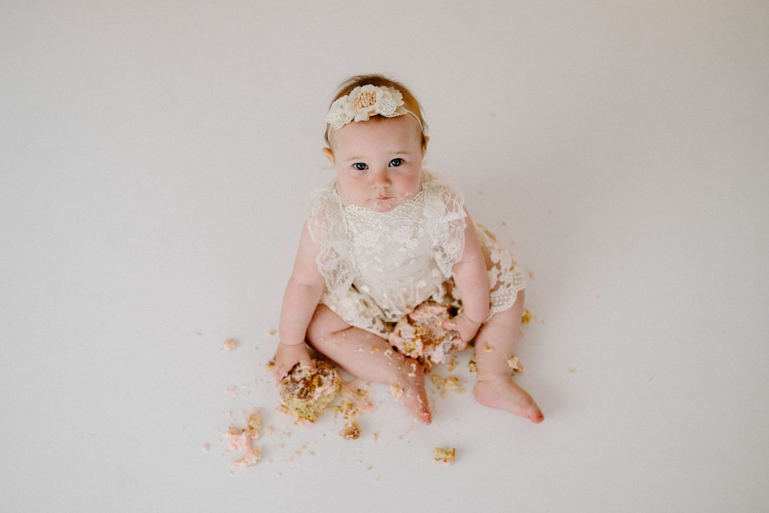 Baby girl in lace romper and gold headband sits amid smashed birthday cake crumbs on white backdrop.