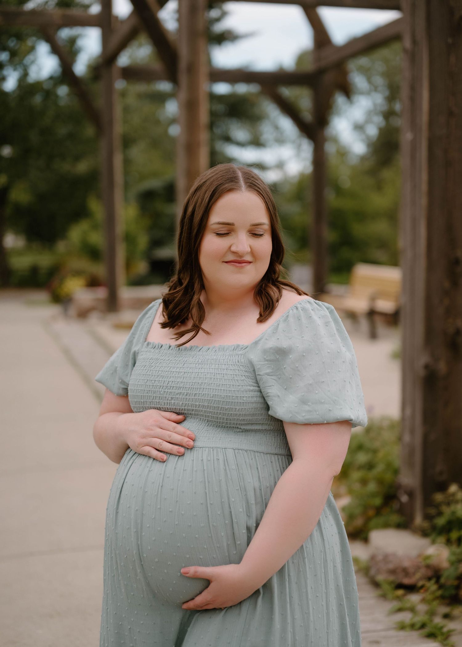 Pregnant woman in sage green smocked dress cradling baby bump outdoors under wooden pergola.