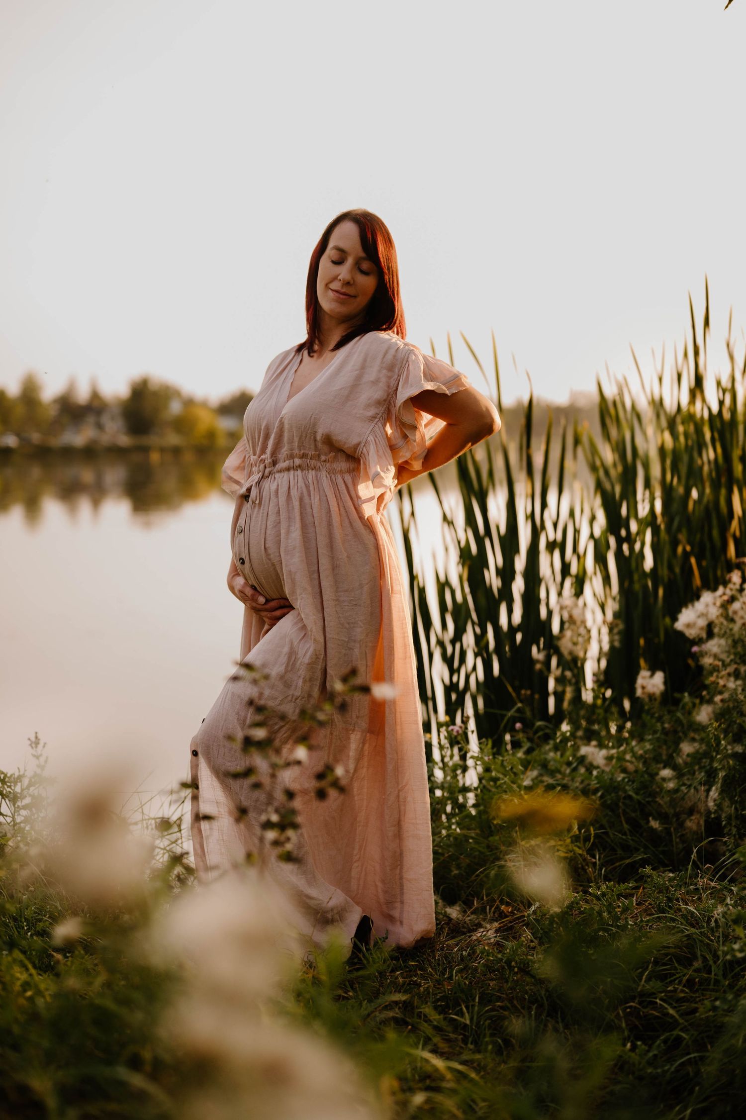 Pregnant redhead woman in pink dress cradling baby bump near lakeside reeds at sunset.