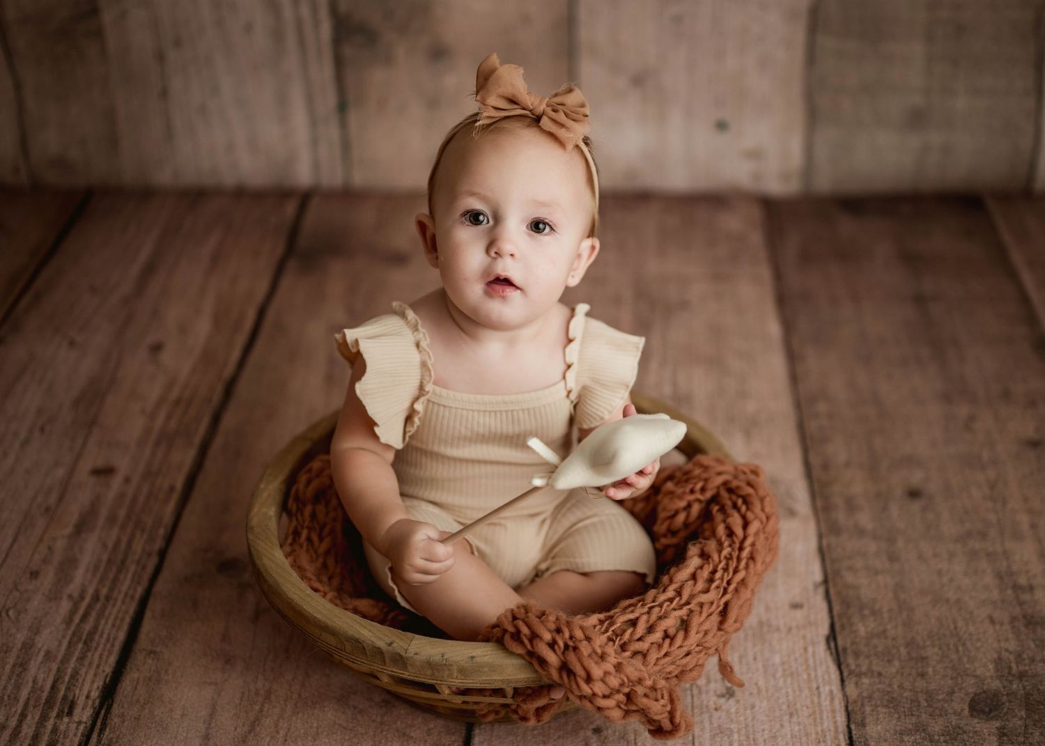 Baby girl in tan romper and bow headband sits in woven basket bowl on rustic wood floor.