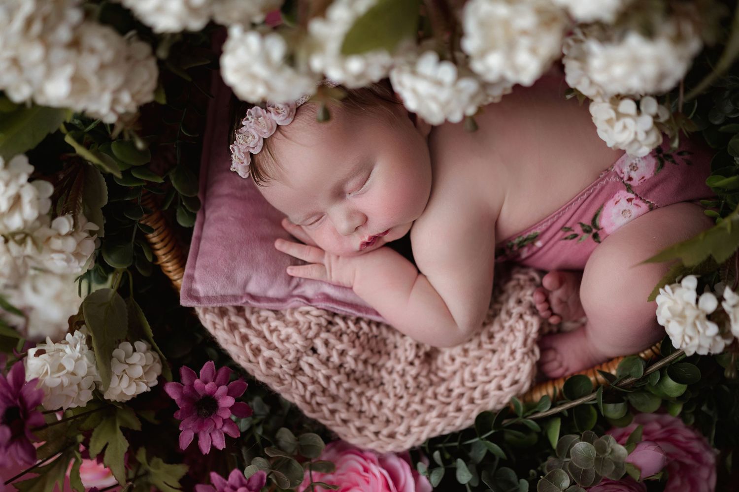Newborn baby girl sleeping peacefully on pink pillow surrounded by white and pink flowers wearing floral headband.