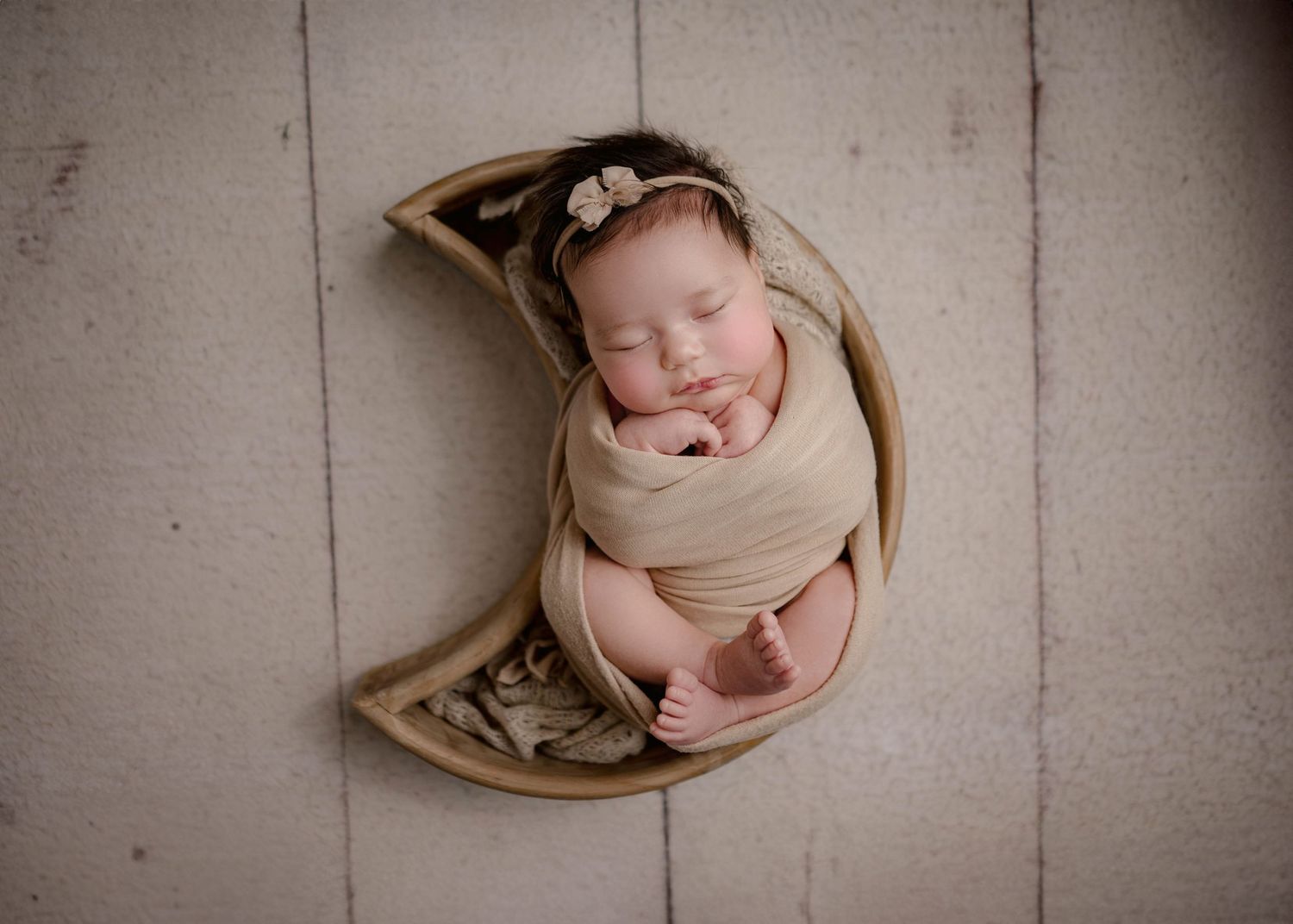 Newborn baby wrapped in beige swaddle sleeping in a crescent moon wooden bowl prop with floral headband.