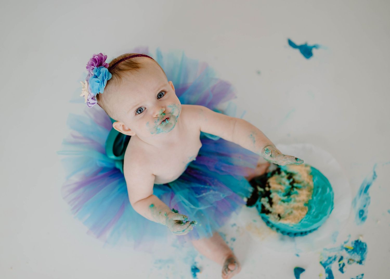 Overhead view of baby girl in colorful tutu during mermaid-themed cake smash session with teal frosting.