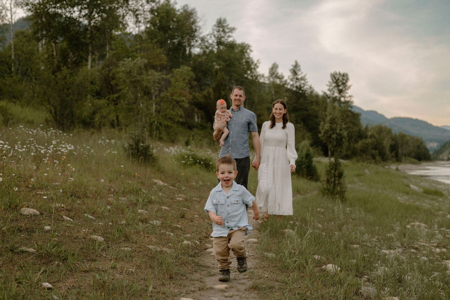 Family of four walking along a scenic riverside trail surrounded by wildflowers and mountains.