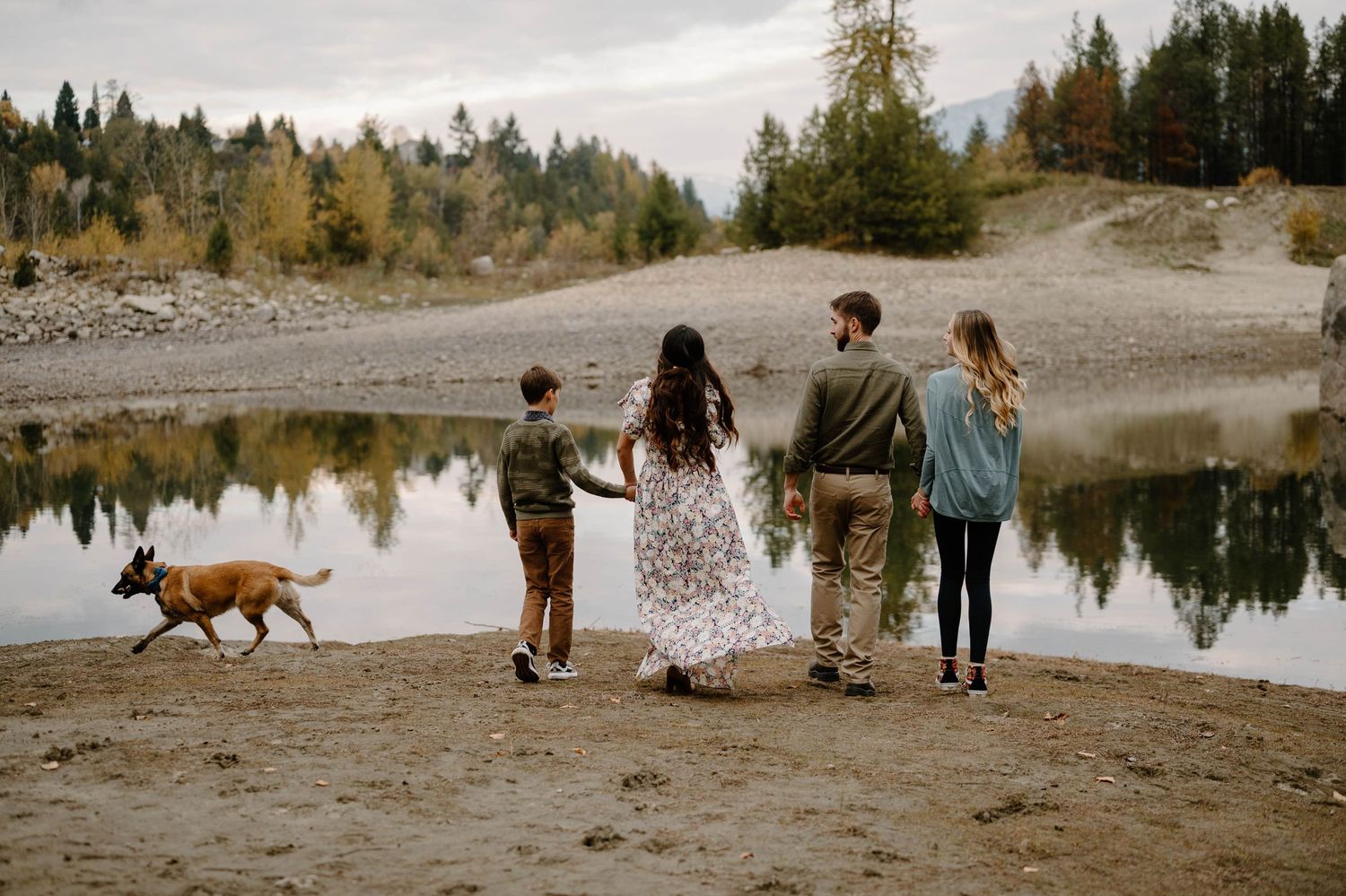 Family with dog standing by a reflective lake surrounded by autumn trees viewed from behind.