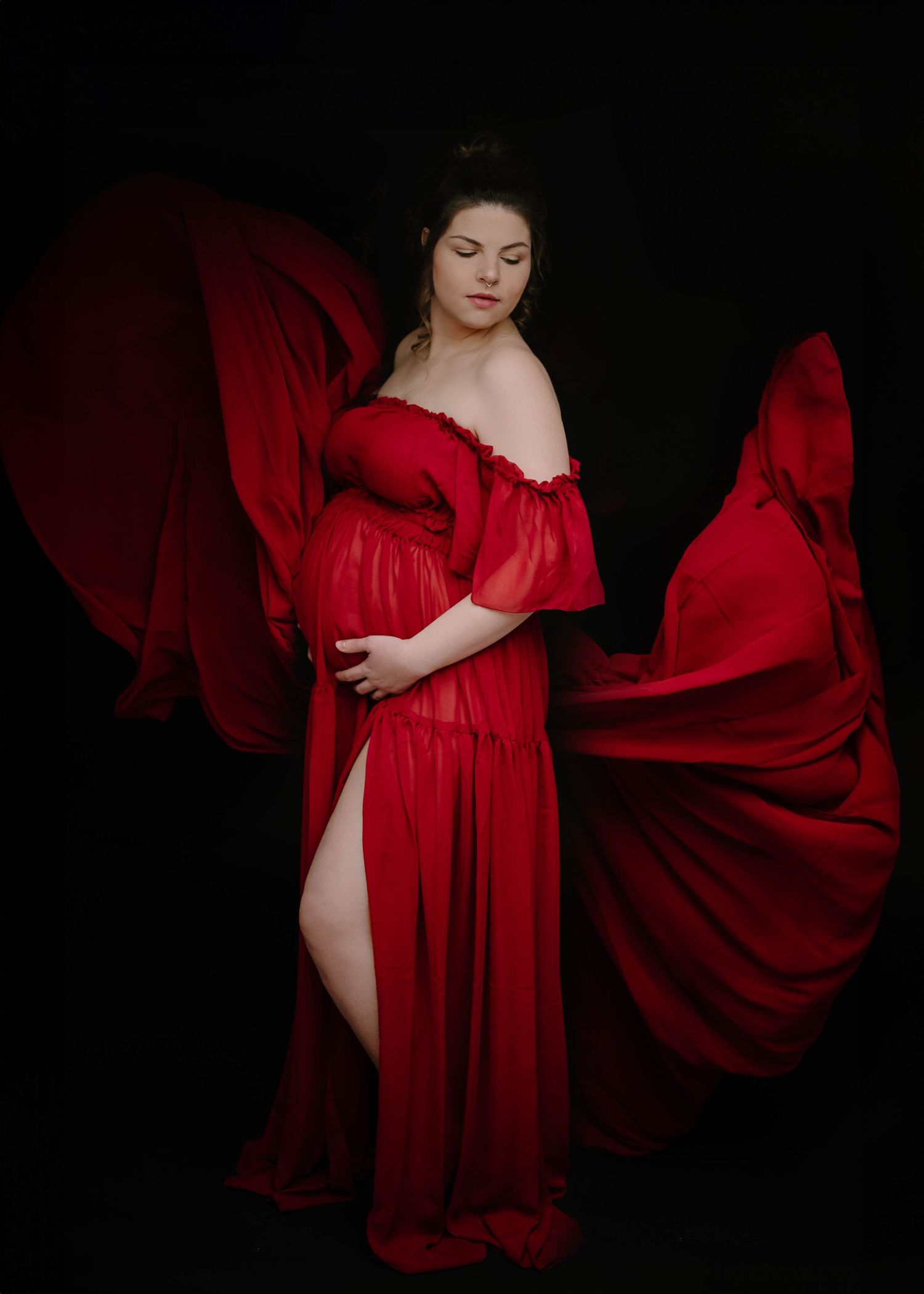 Pregnant woman in flowing red off-shoulder gown with fabric wings on black background.