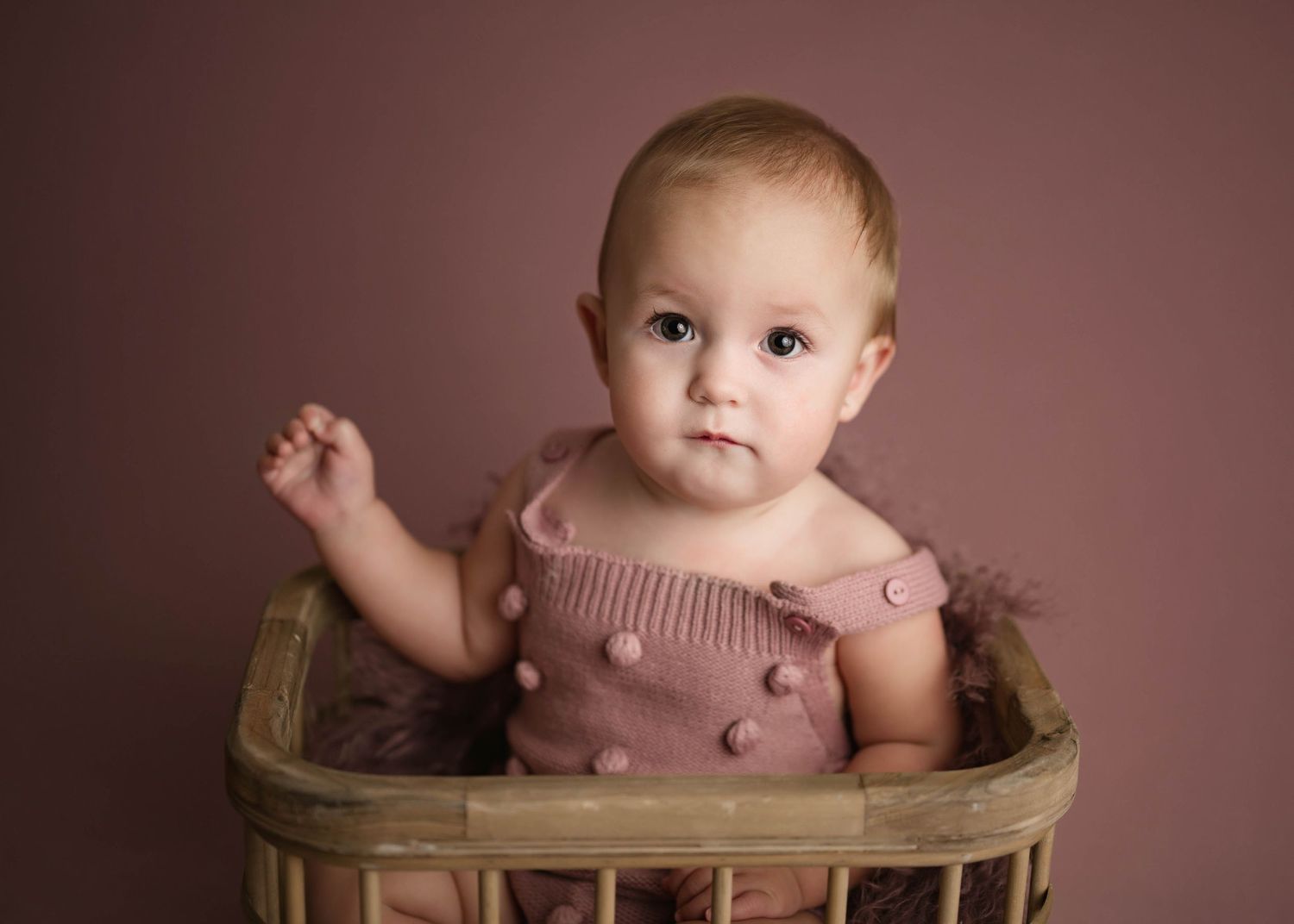 Baby girl in a mauve knit romper sitting in a rustic wicker basket against a pink studio backdrop.