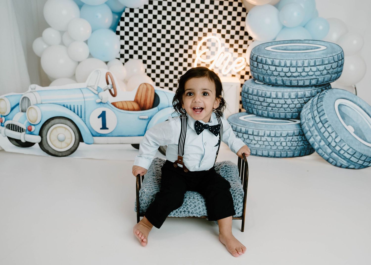 Happy toddler boy sits in a vintage race car themed first birthday cake smash photo setup.