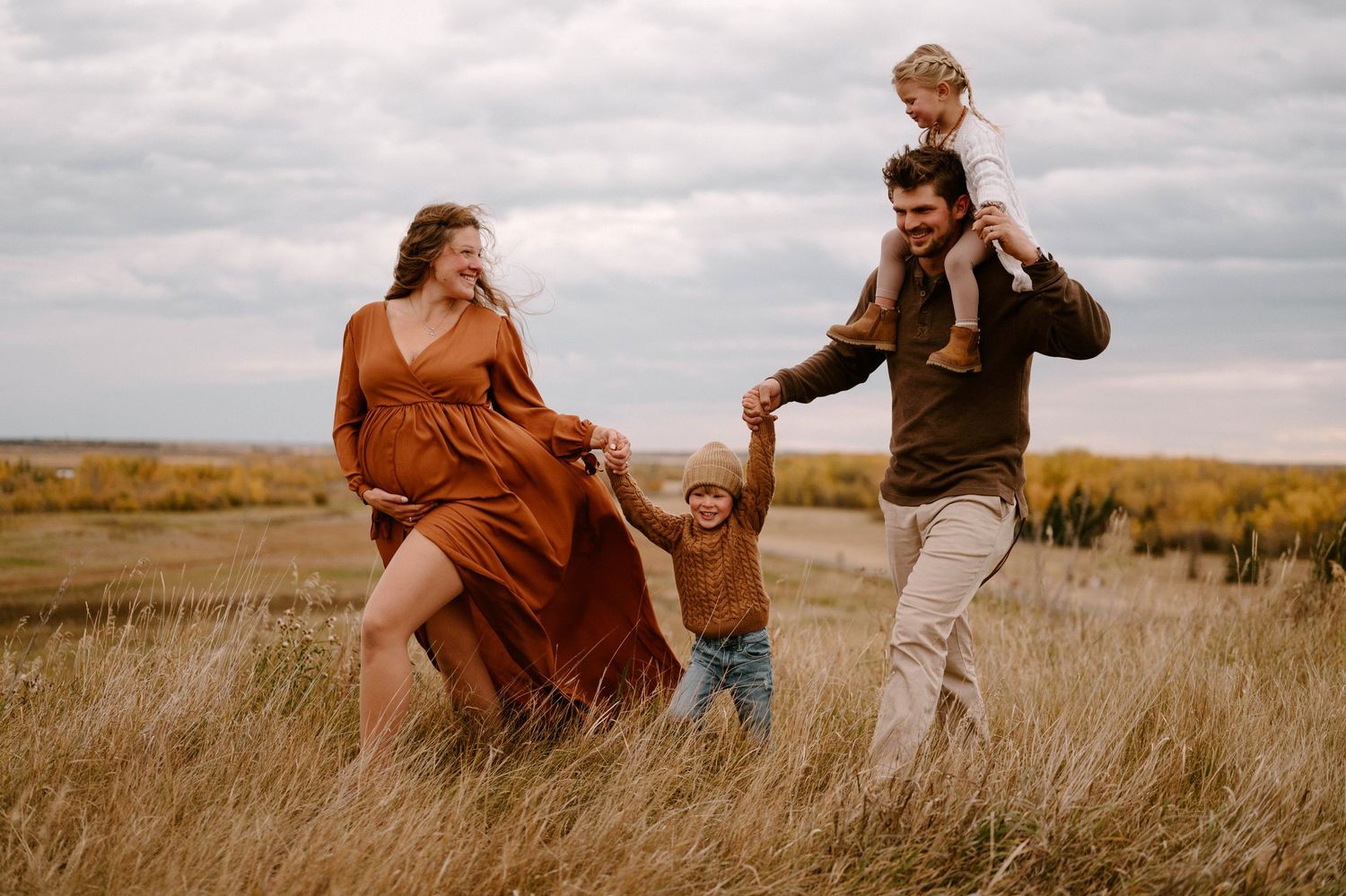 Happy family walking in golden autumn field, pregnant mother in rust dress holding hands with children.