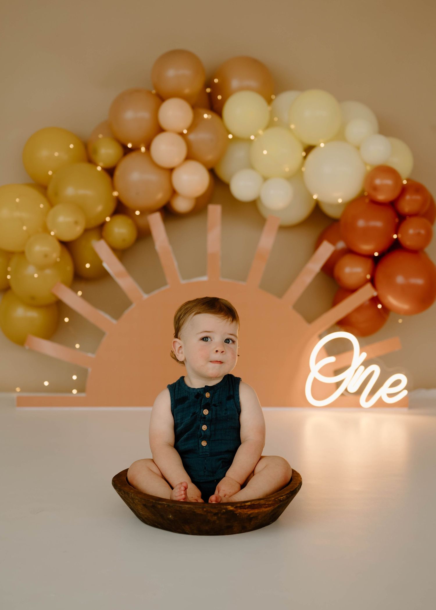 Cute baby boy in teal romper sitting in wooden bowl beneath boho sun balloon arch decor.