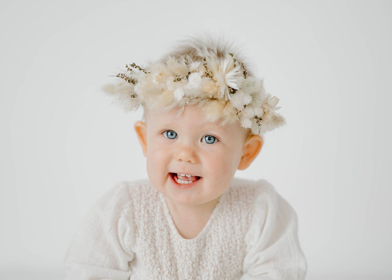 Adorable smiling baby girl with blue eyes wearing white floral crown and white knit outfit.