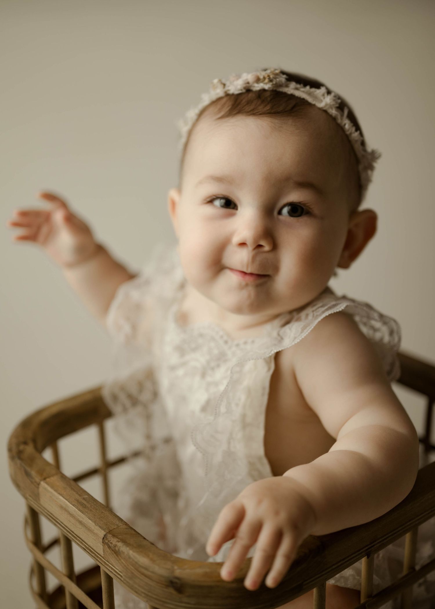 Happy baby girl in white lace outfit standing in rustic wicker basket with arm raised joyfully.