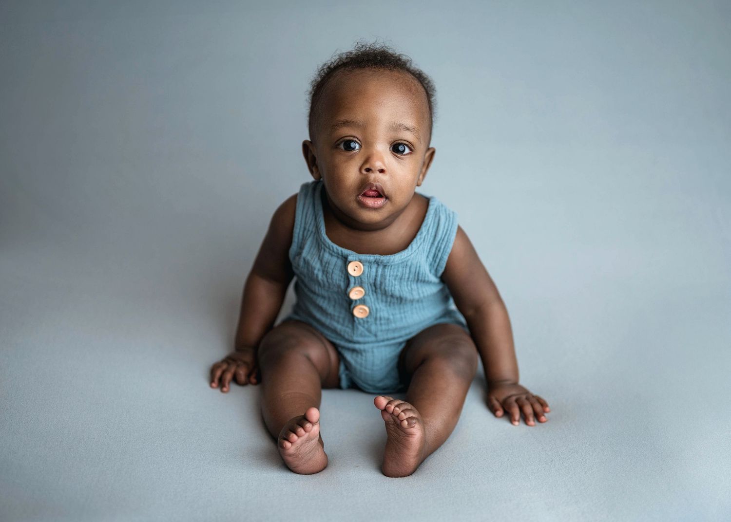 Adorable baby in blue romper sitting upright on gray background.