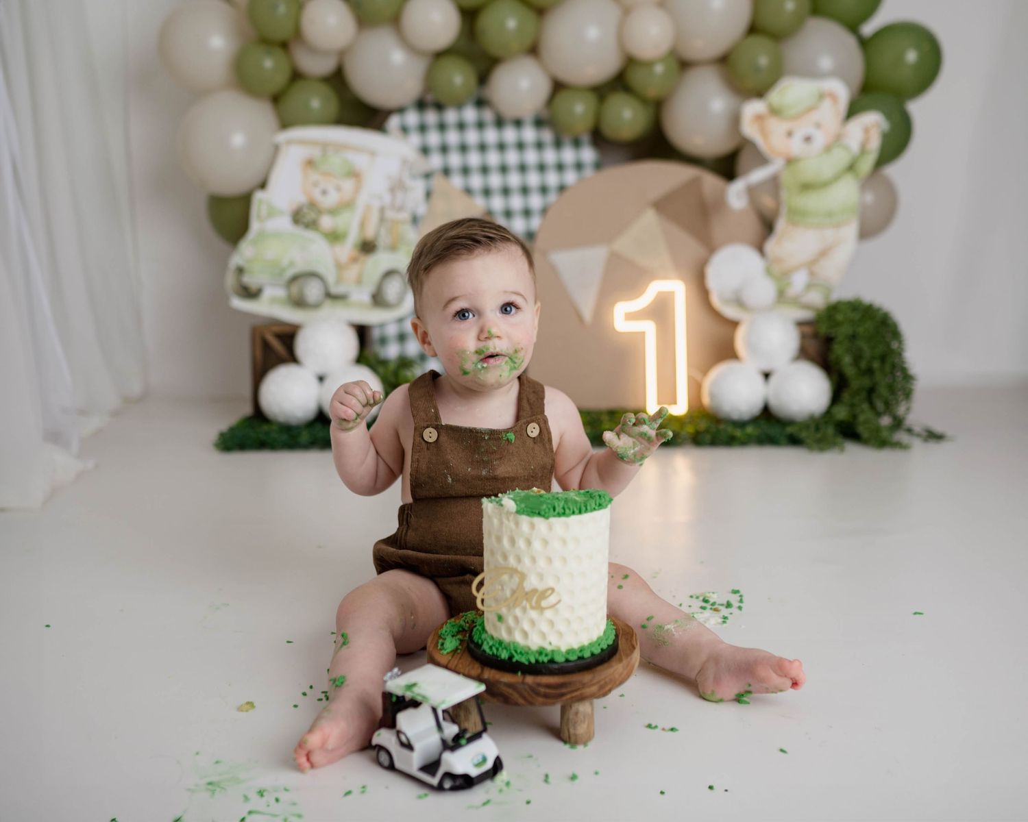 Baby's first birthday cake smash with golf theme, green balloons, number 1 sign, and chocolate cake on white backdrop.