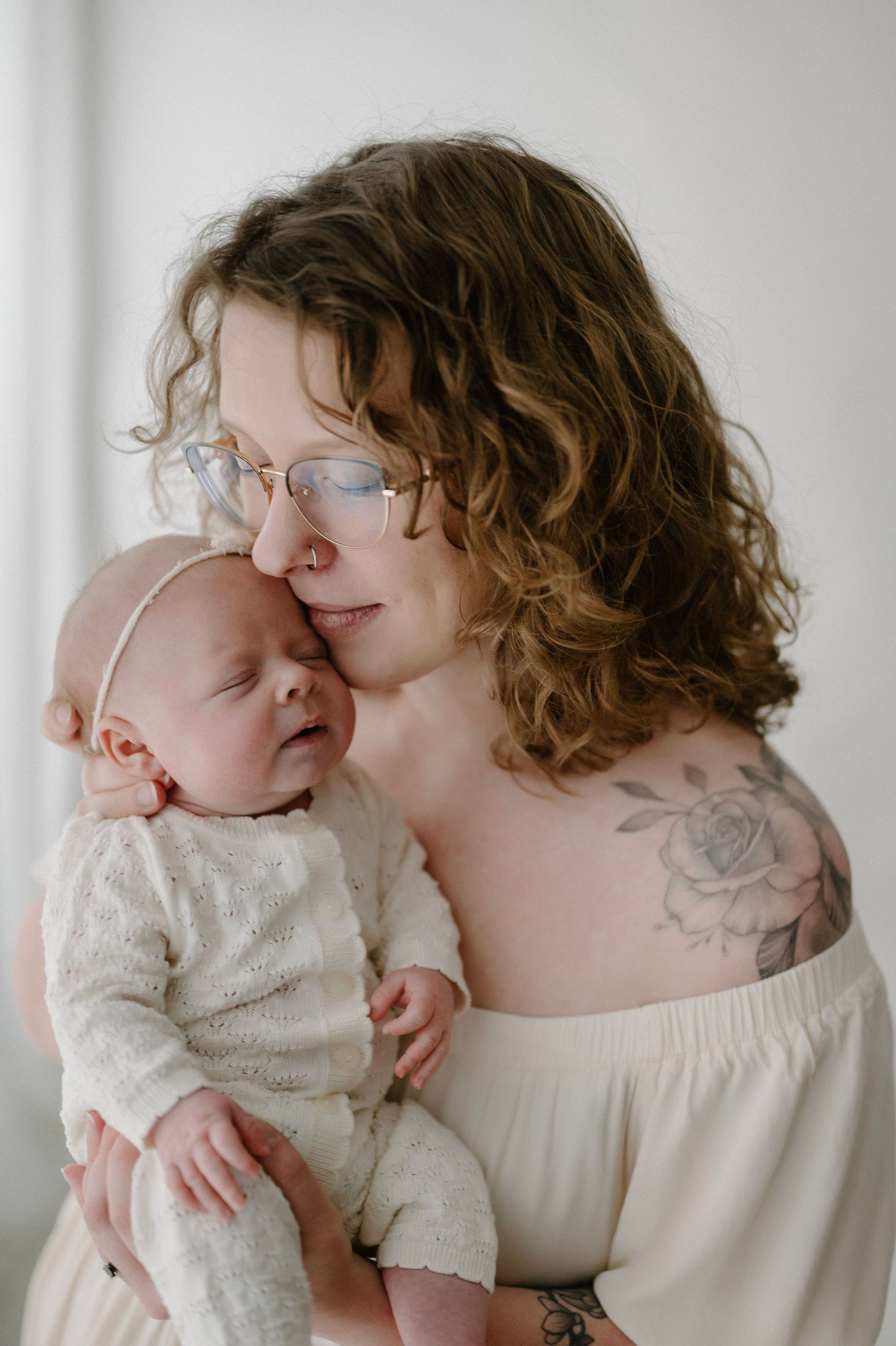 A curly-haired mother with glasses and floral tattoo tenderly holds her sleeping newborn baby close to her cheek.