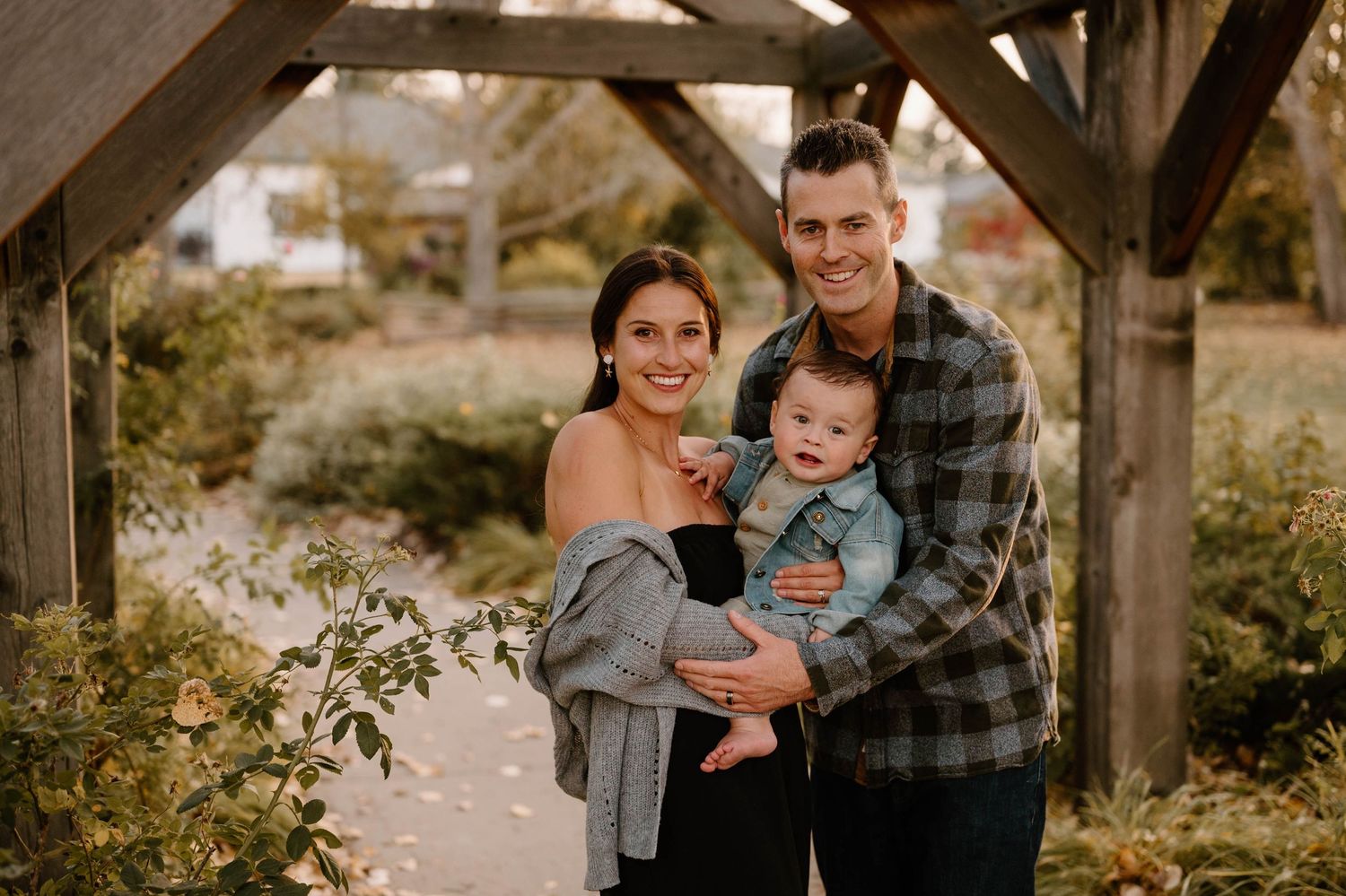 Happy family of three posing under wooden pergola in autumn park, parents smiling with baby boy.