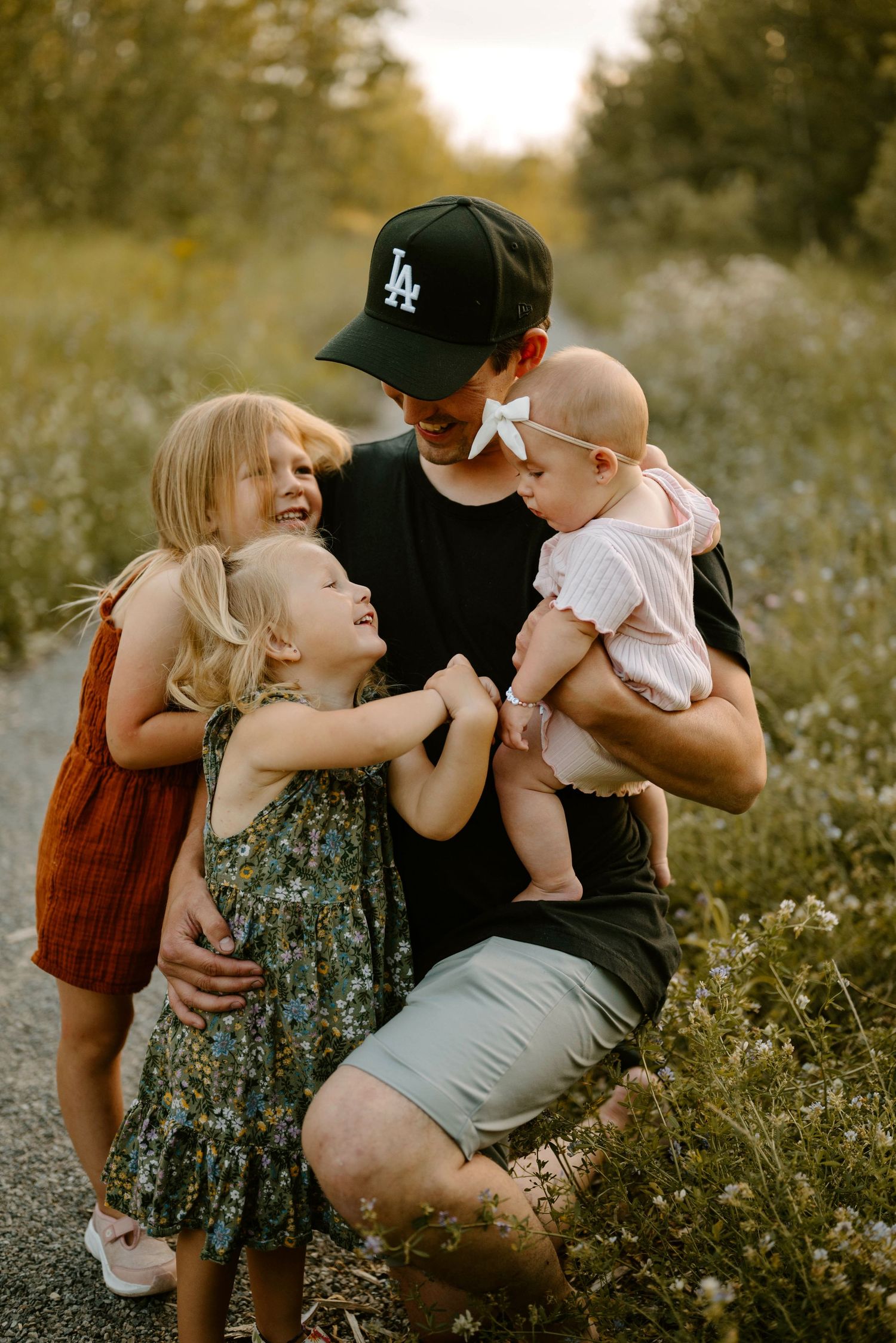 Father kneeling on a path laughing with three young daughters in golden hour light.