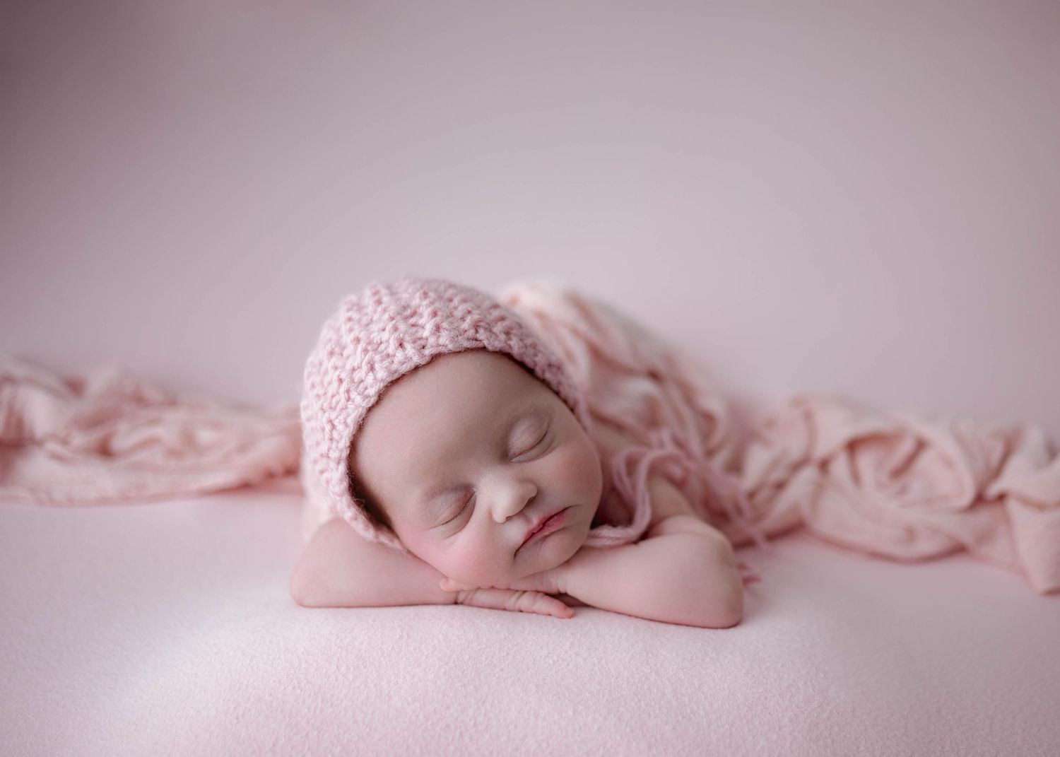 Peaceful newborn baby sleeping in pink knit bonnet on soft pink blanket in professional newborn photography portrait.