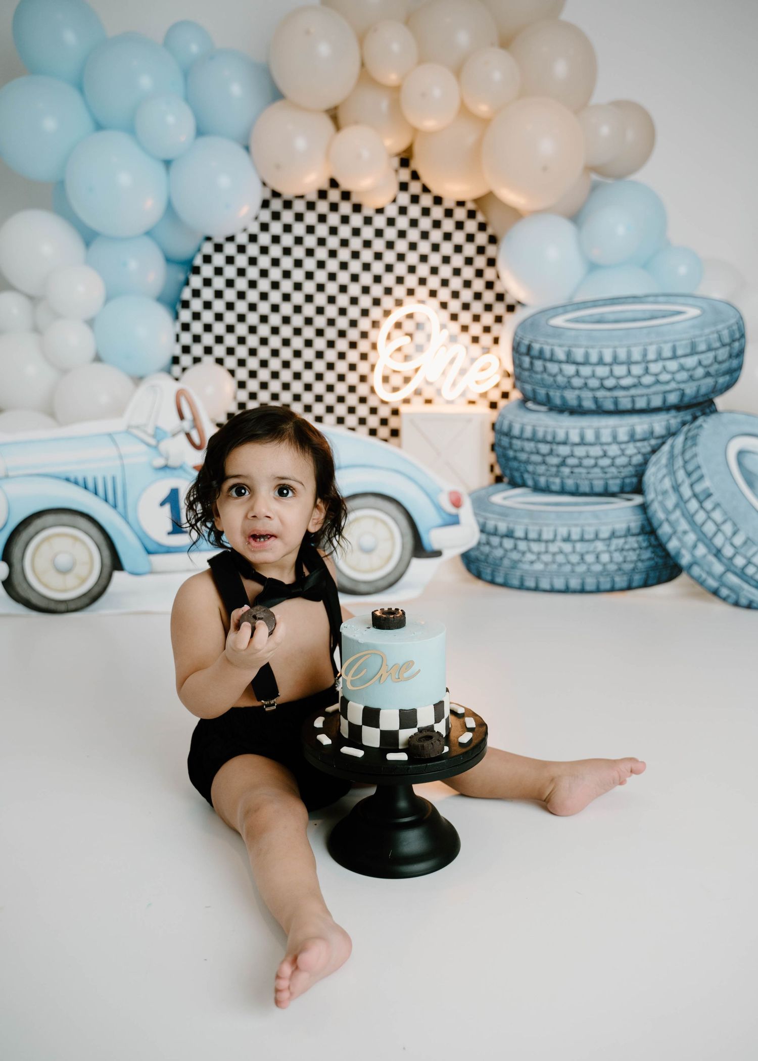 A baby boy in a black romper sits with a checkered race car themed birthday cake during a first birthday cake smash session.