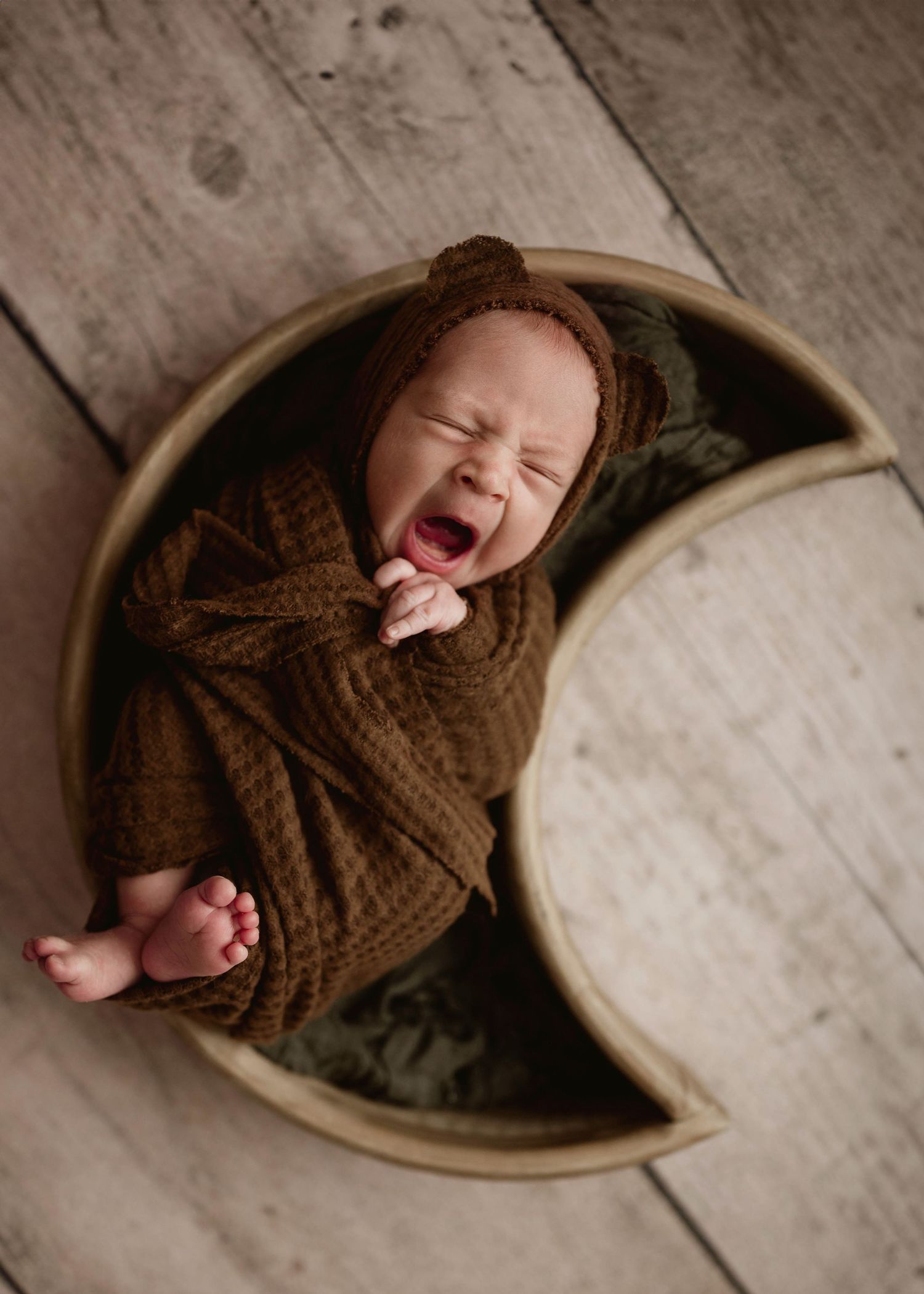 Yawning newborn baby wrapped in brown swaddle wearing bear bonnet in crescent moon shaped prop.