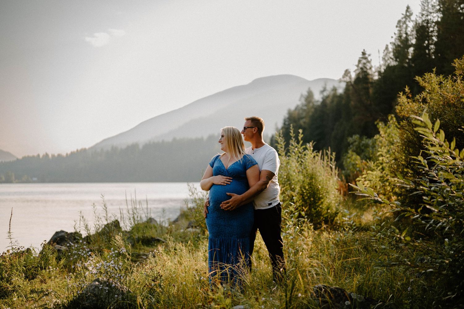 Maternity couple embracing by mountain lake surrounded by lush green foliage.