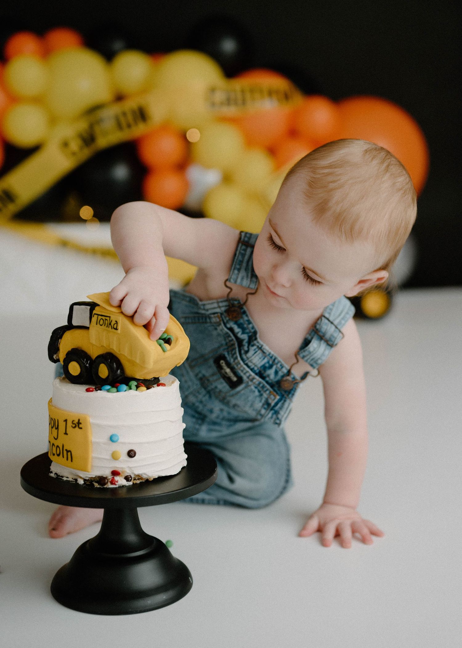 Baby boy in denim overalls grabs toy dump truck from decorated first birthday cake on black stand.