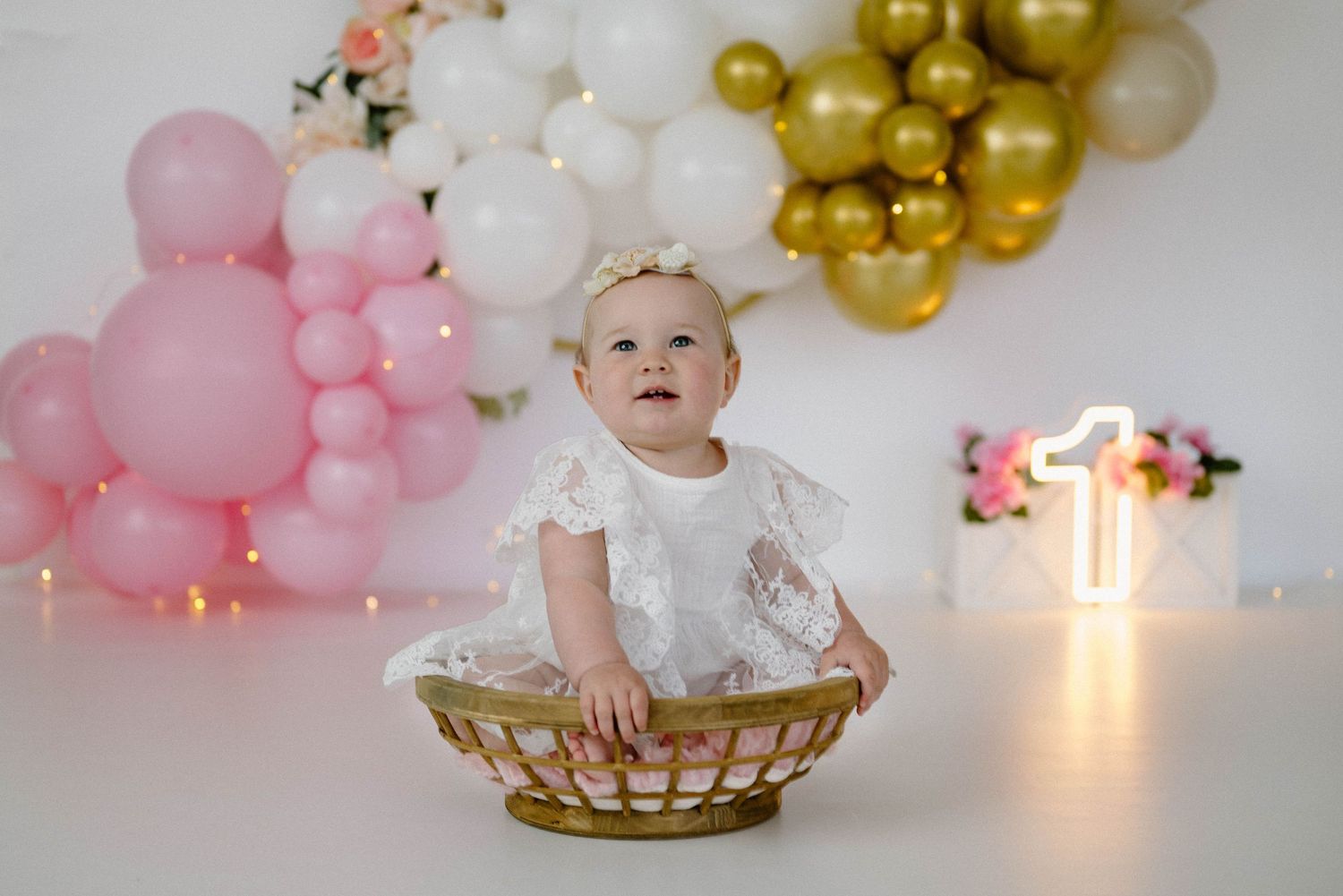 Baby girl in white lace dress sits in wicker basket at first birthday with pink and gold balloon garland.