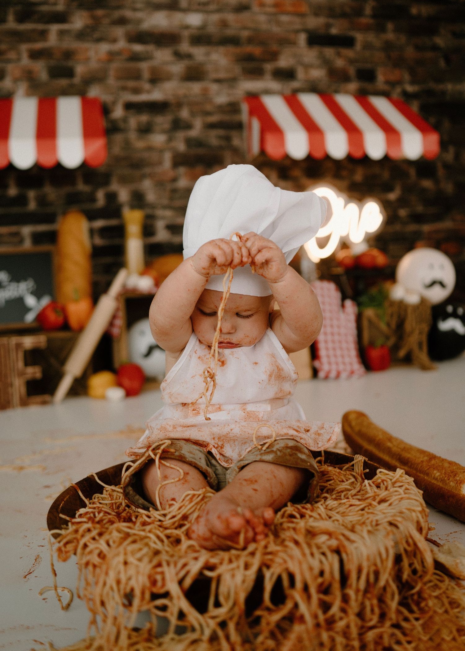 Baby in chef hat plays with spaghetti in bowl during Italian-themed first birthday smash session.