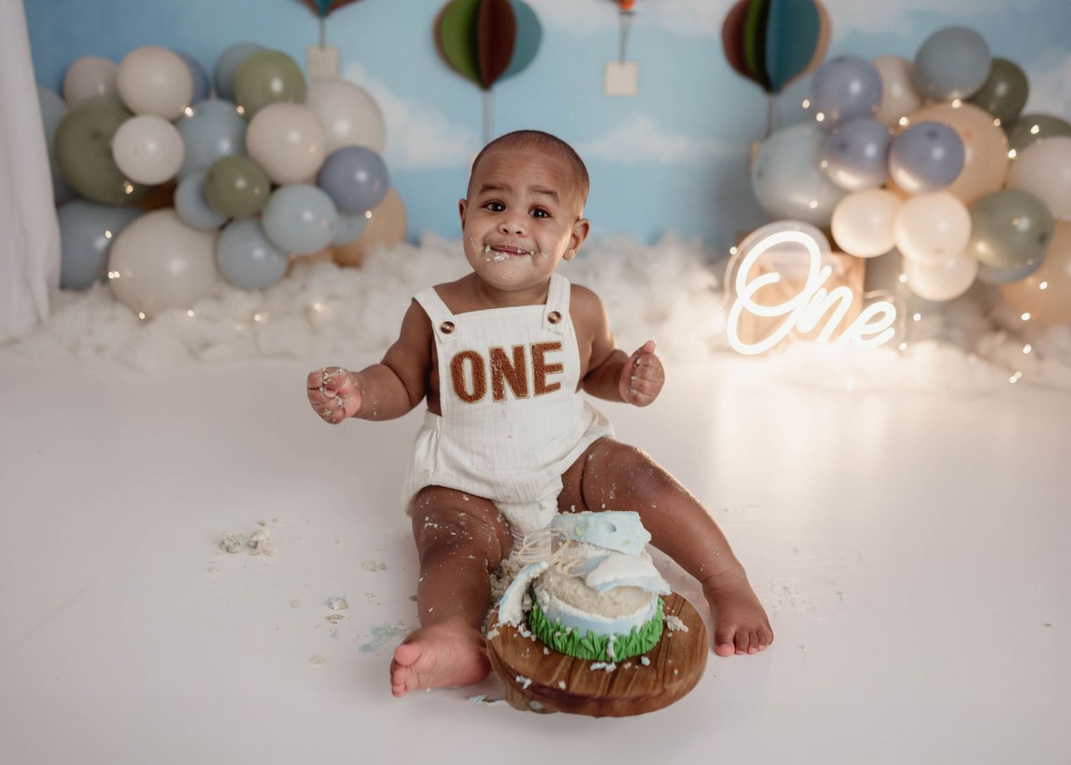Messy baby boy covered in cake at a hot air balloon first birthday cake smash session with neon One sign backdrop.
