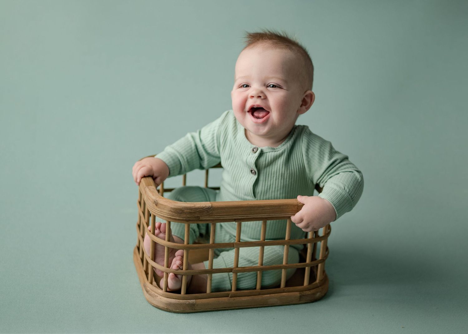 Laughing baby boy in sage green romper with mouth wide open sitting in a wicker basket.