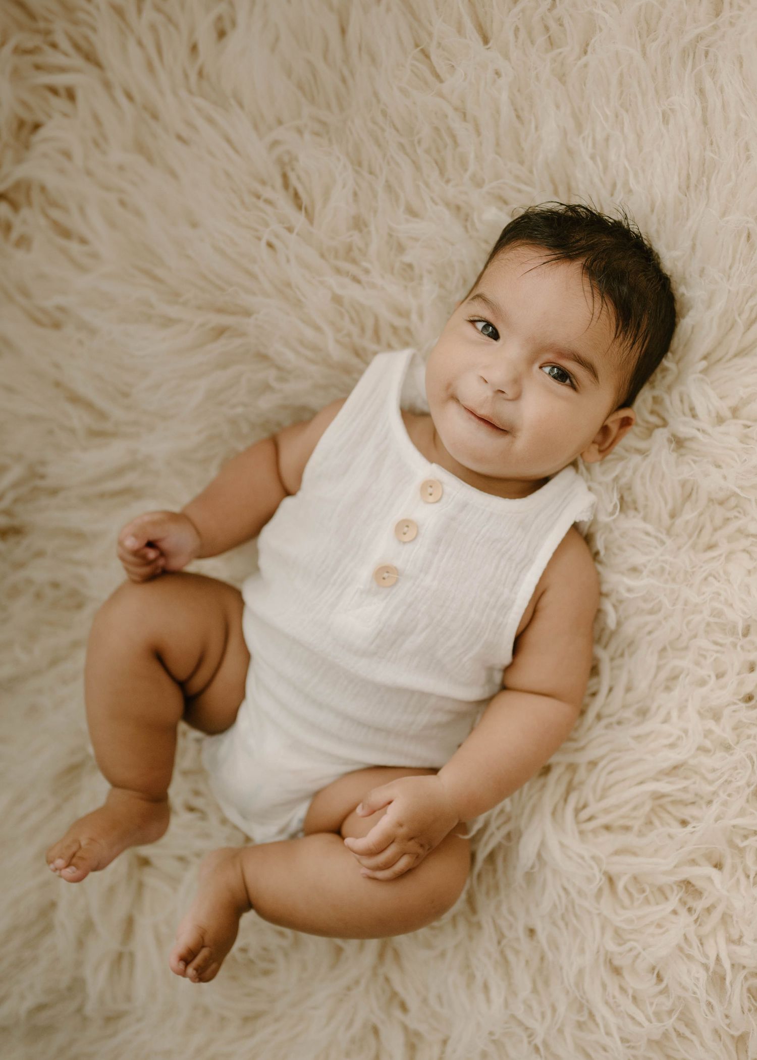 Smiling baby in a white sleeveless romper reclining on a fluffy cream rug, looking up at the camera.