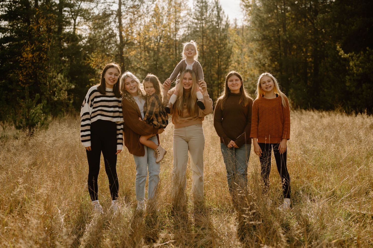 Seven girls pose together in a golden autumn field surrounded by tall grass and trees.