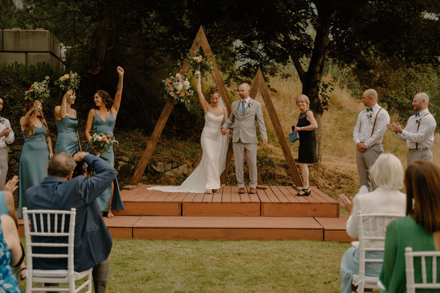 Bride and groom raising hands in celebration at an outdoor ceremony with guests and a triangular wooden arch.