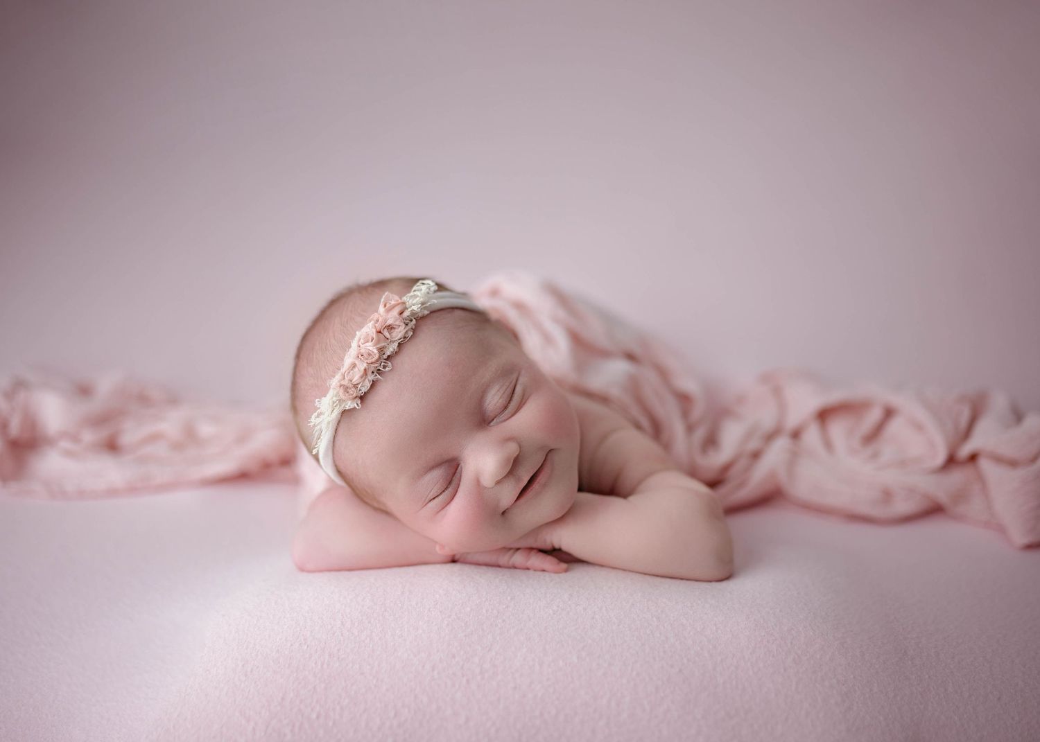 Smiling sleeping newborn baby girl with floral headband posed on soft pink blanket backdrop In home Leduc studio.