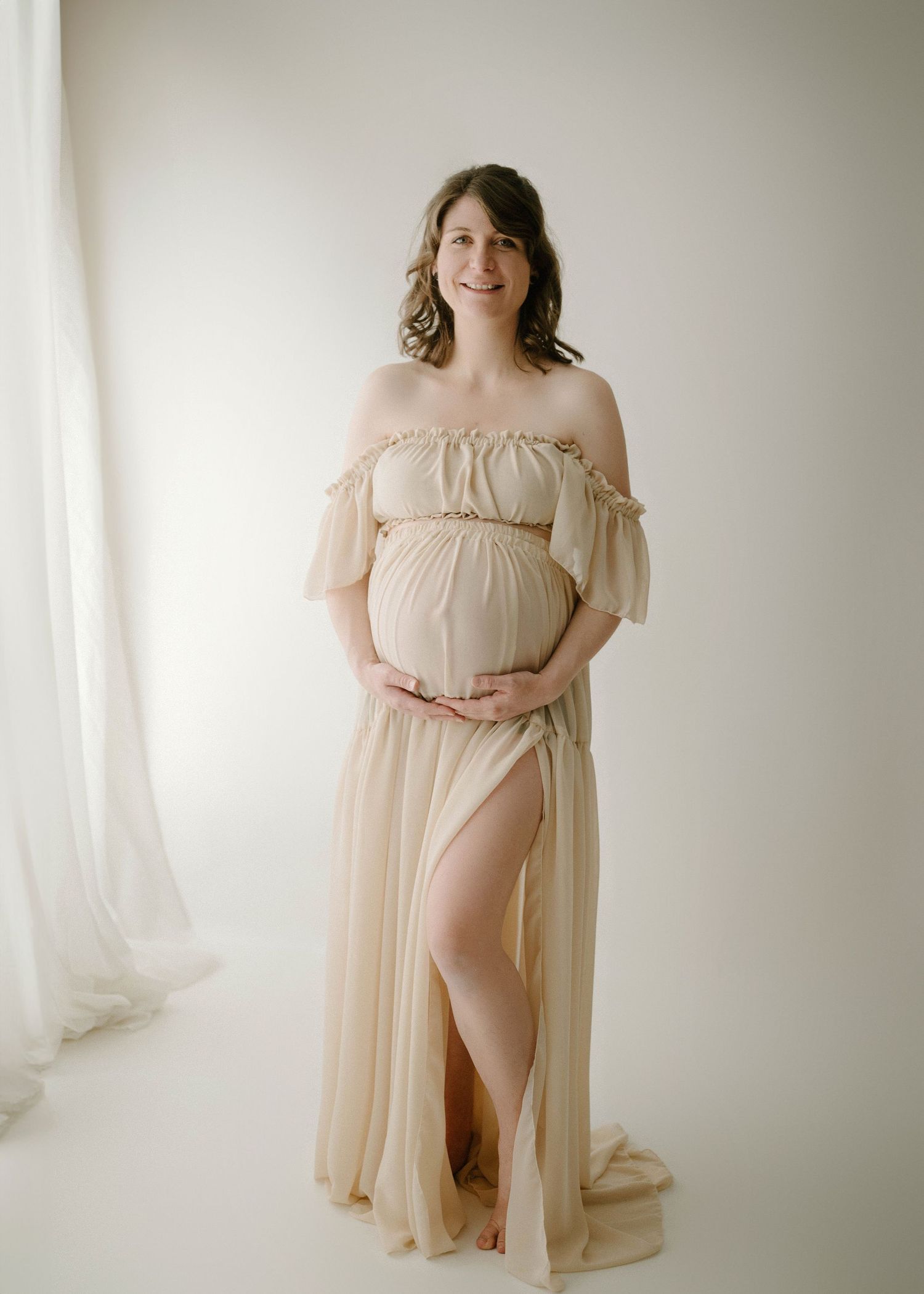 Radiant pregnant woman in flowing off-shoulder champagne gown holds belly in bright studio.