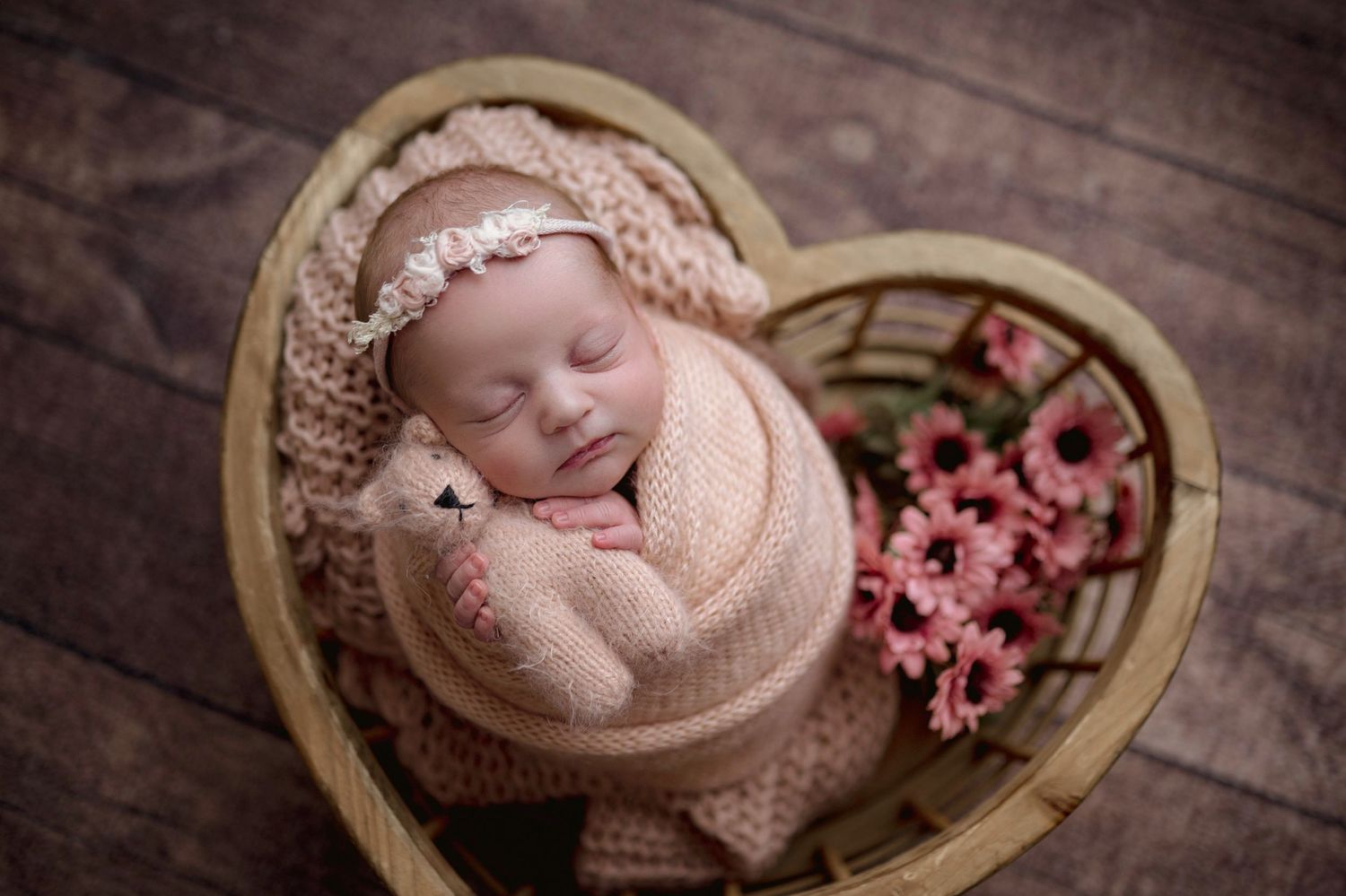 Newborn baby sleeping peacefully in a heart-shaped basket with pink flowers and a floral headband.