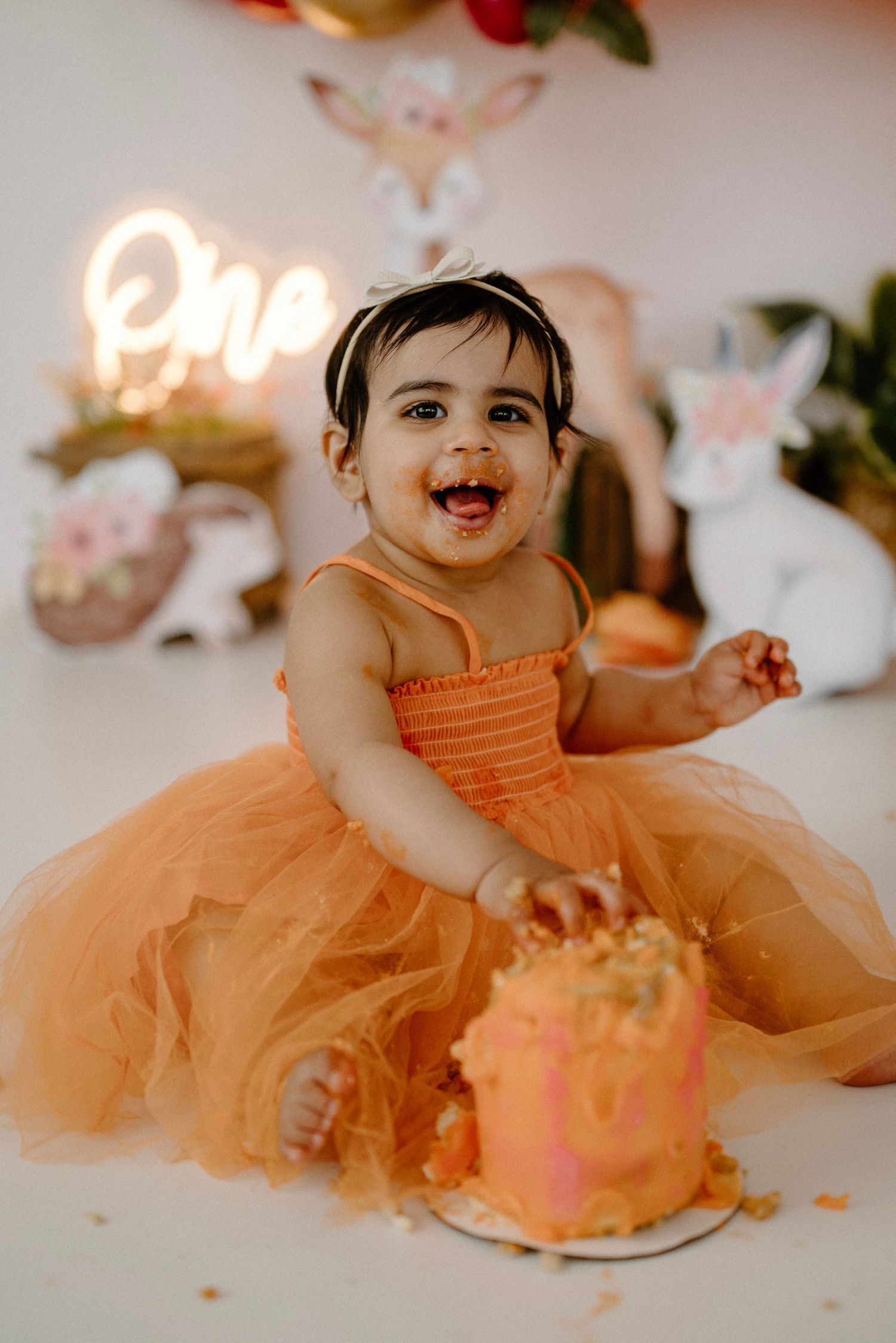 Happy baby girl in orange tutu laughing while smashing a pink birthday cake at party.
