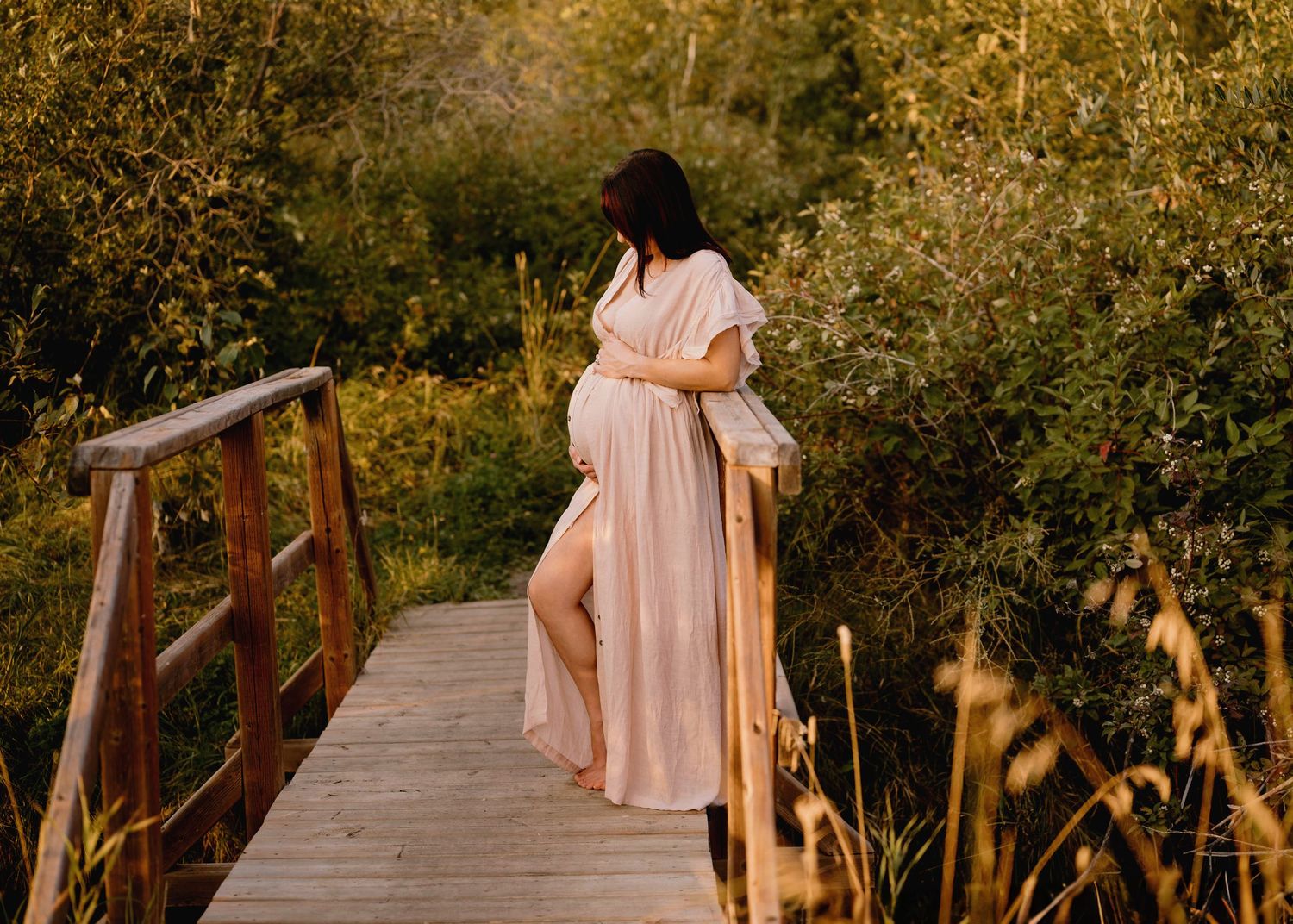 Pregnant woman in blush maxi dress walks across wooden bridge at Telford Lake in Leduc surrounded by golden autumn foliage.