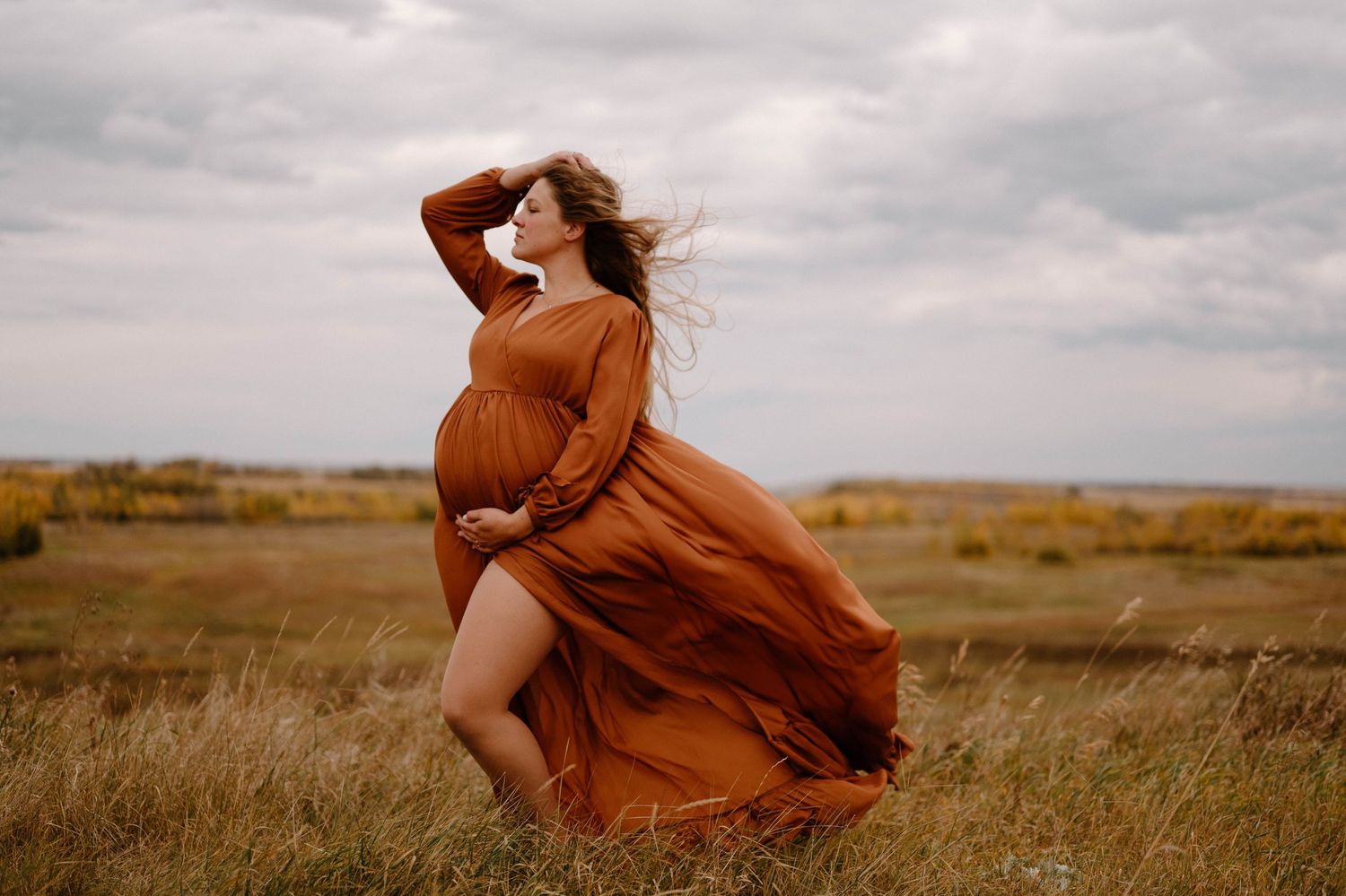 A pregnant woman in a flowing rust dress poses dramatically in a windswept autumn field.