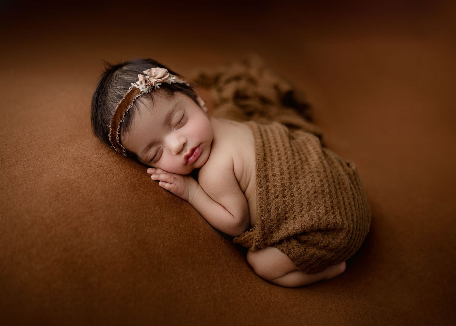 Sleeping newborn baby wrapped in brown knit blanket with floral headband on brown background.