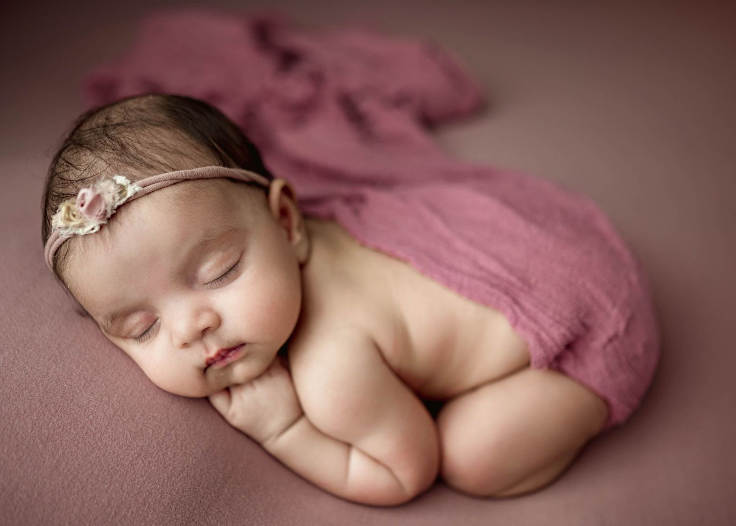 Peaceful newborn baby girl sleeps curled up on a mauve pink blanket wearing a delicate floral headband.