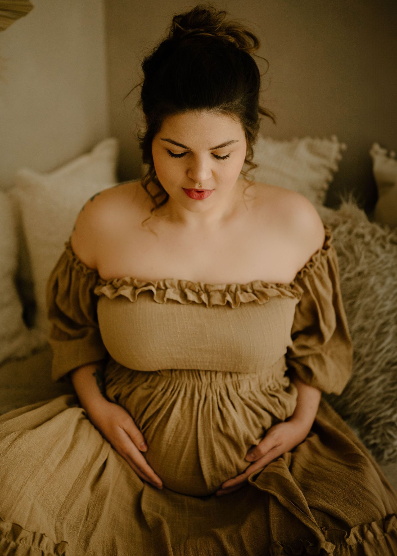 Pregnant woman in golden off-shoulder dress gently holding baby bump on bed.