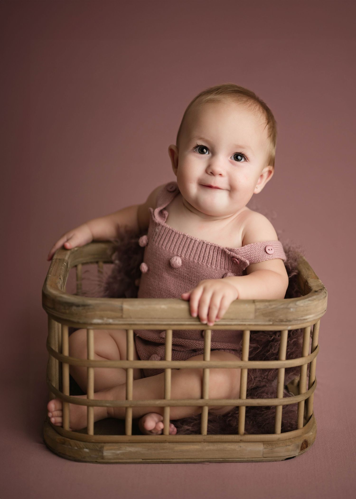 Cheerful baby girl in a mauve knit romper posing in a wicker basket against a dusty rose backdrop.