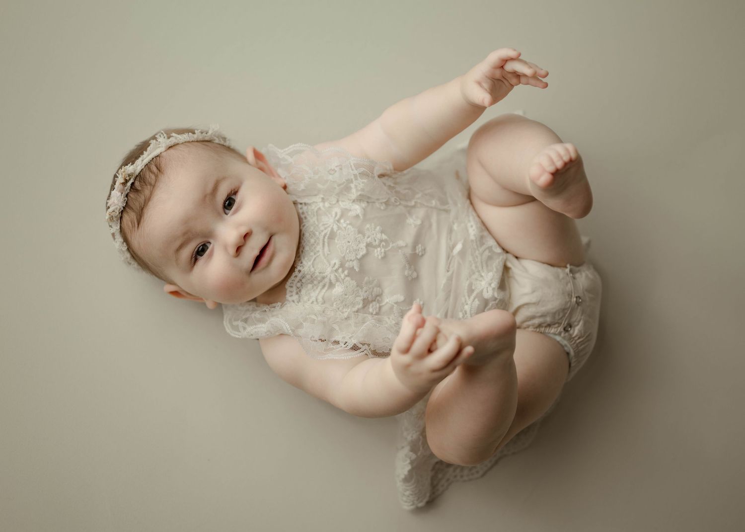 Baby girl in white lace romper and pearl headband lying on cream backdrop during professional studio session.