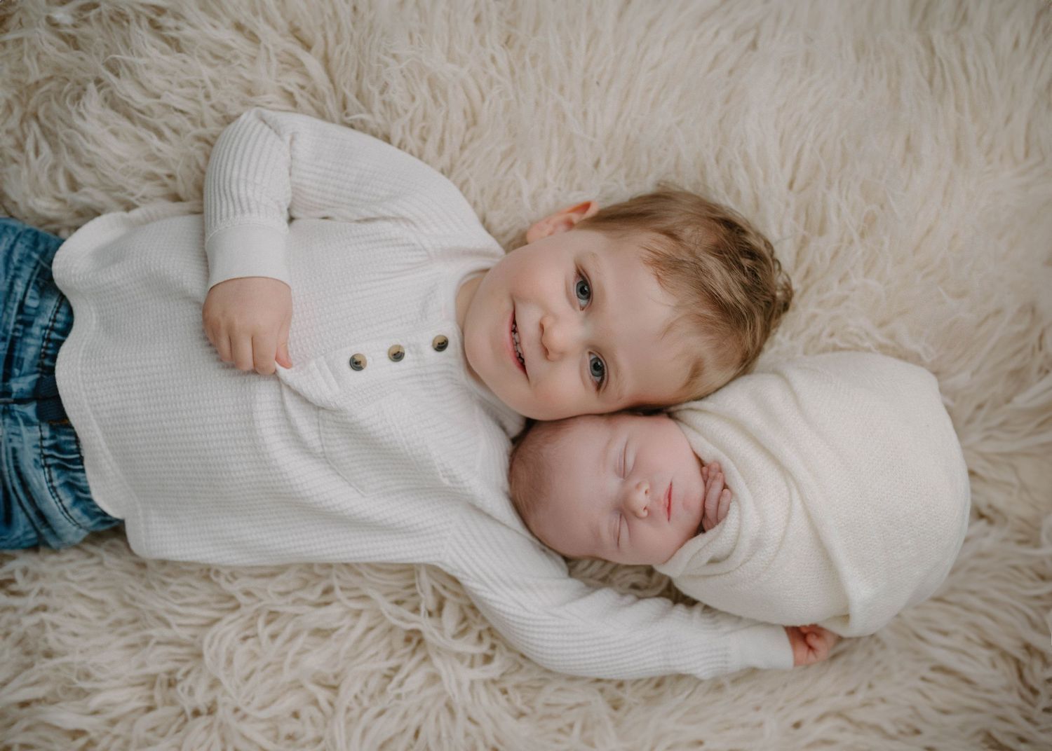 Toddler boy in white sweater lying on fluffy rug with sleeping newborn baby wrapped in white swaddle.